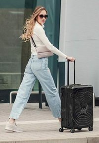 Černý tvrdý kufr s vlnkovým vzorem, teleskopická rukojeť a kolečka. Neformální outfit: bílý svetr, světle modré džíny a tenisky.