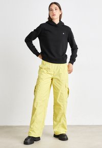 Czarna bluza z kapturem, żółte spodnie cargo z bocznymi kieszeniami, masywne czarne buty na platformie, gładki materiał i luźny krój.