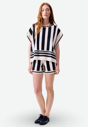 Jeune femme portant un haut en maille à rayures verticales noires et blanches assorti à un short, avec des baskets noires et un collier de perles, debout droit.