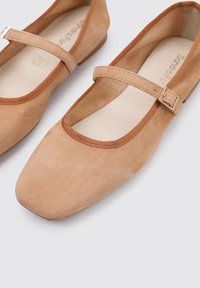 Ballerines en daim tan avec un bout carré, présentant un bord arrondi et une seule sangle avec une boucle métallique. Doublure intérieure lisse, design minimaliste.