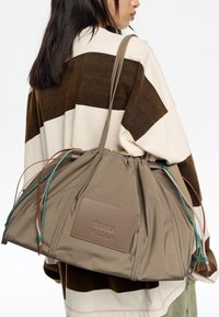 Bolso de tela marrón y beige con cierre de cordón, parche de cuero y acentos de cordón multicolor. Textura suave, forma redondeada.