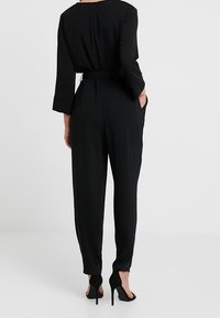 Svart jumpsuit med långa ärmar, bälte i midjan och fickor. Har rund halsringning och tapered ben. Materialet är slätt och enhetligt.