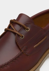 Sapato de barco em pele castanha com atacadores castanhos claros, costura detalhada e sola de borracha, com uma marca subtil em baixo-relevo na lateral.