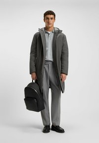 Grauer langer Mantel mit Kapuze, kombiniert mit einem hellgrauen Poloshirt und grauen Hosen. Trägt einen schwarzen strukturierten Rucksack und schwarze Schuhe.