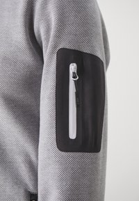 Gray textured fabric sweater with a black patch at the upper arm, featuring a white zippered pocket and a small logo tag.