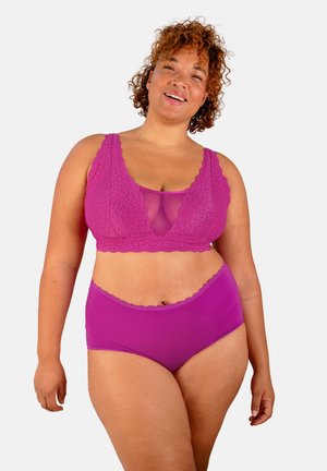 Smiling woman with curly hair wearing matching purple lace bralette and high-waisted underwear, standing against a plain background.