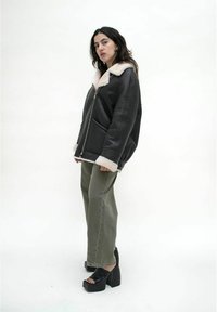 Schwarze Lederjacke mit einem weißen Kunstfellkragen, Reißverschluss und seitlichen Taschen. Getragen mit olivgrünen, weit geschnittenen Hosen und schwarzen Plateauschuhen.