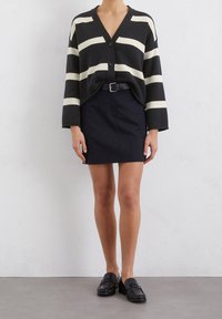 Schwarz-weißer gestreifter Cardigan mit V-Ausschnitt und Knopfverschluss, kombiniert mit einem taillierten schwarzen Rock und schwarzen Slippern. Matte Stofftextur.