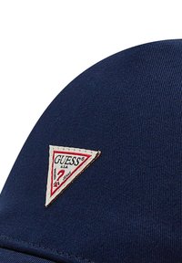 Mörkblå keps med en triangulär vit och röd logotypapplikation som har texten "GUESS", tillverkad av texturerat bomullsmaterial, med en böjd skärm.