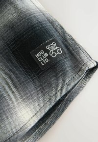 Denimstof i sort og grå gradient med en sort label, der har en hvid bjørnegrafik og teksten "HUG CLUB LTD." Glat tekstur.