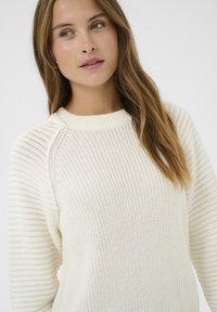 Jeune femme aux longs cheveux châtain clair portant un pull en tricot texturé blanc, regardant légèrement sur le côté devant un fond uni.