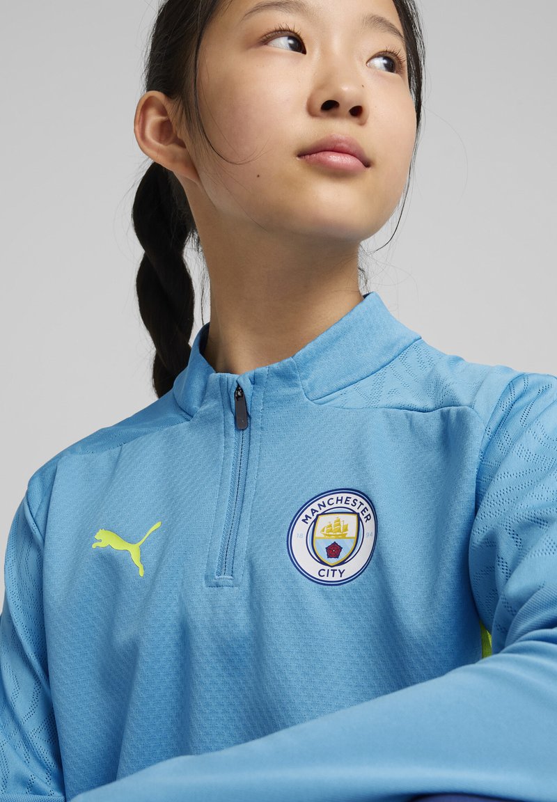 Veste de sport bleu ciel avec col zippé, tissu texturé et logo Puma jaune. Présente le blason du club de Manchester City sur la poitrine.