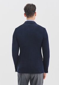Granatowa kurtka o dopasowanym kroju, z wyraźnie zarysowanymi ramionami i eleganckim designem. Gładka tekstura z dwoma wentylami z tyłu i mankietami zapinanymi na guziki.