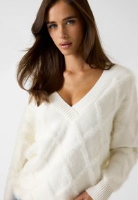 Maglione bianco morbido con scollo a V e polsini a coste, caratterizzato da un motivo a diamante testurizzato e una vestibilità ampia. Tessuto morbido e caldo.