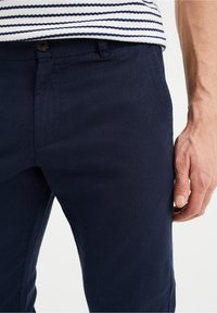 Man som bär marinblå byxor och en vit skjorta med marinblå horisontella ränder, hand vilande vid sidan.