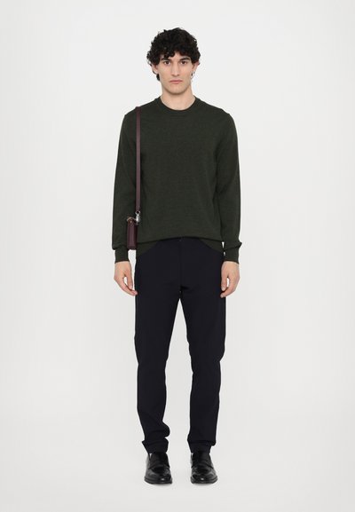 Dunkelgrüner gestrickter Pullover, leichte Textur; marineblaue Hose; schwarze Schuhe; braune Umhängetasche; Modell steht vor einem schlichten weißen Hintergrund.