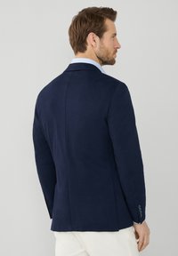 Blazer navy con rever a punta, apertura centrale e chiusura a tre bottoni. Presenta una texture liscia e dettagli del collare azzurro chiaro.