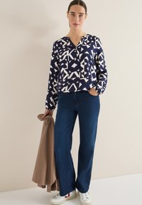 Marineblauwe blouse met een patroon van witte geometrische ontwerpen, lange mouwen en een v-hals. Gepaard met hoge blauwe jeans en witte sneakers.