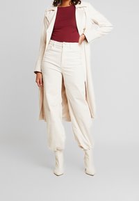 Pantaloni in velluto a coste beige con una vestibilità rilassata, abbinati a un top bordeaux e a un lungo cappotto crema. L'outfit è completato da stivali bianchi alla caviglia.