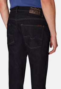 Jeans in denim scuro con cuciture a contrasto, due tasche posteriori e un'etichetta in pelle marrone testurizzata sulla vita. Vestibilità regolare, minimale usura.