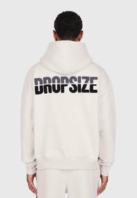 Person wearing white hoodie with large black and gray "DROPSIZE" text on the back, standing against a plain light background.