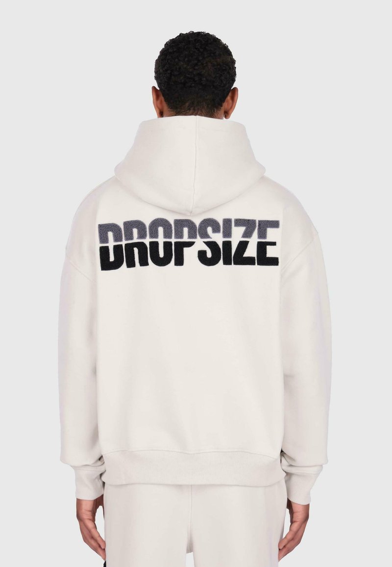 Person wearing white hoodie with large black and gray "DROPSIZE" text on the back, standing against a plain light background.