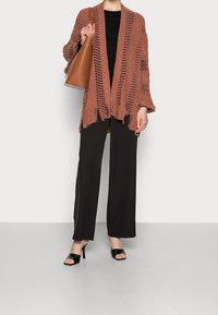 Rdzawoczerwony, dziany kardigan z luźnym splotem i frędzlami na dolnym końcu, zestawiony z czarnymi spodniami w szerokim kroju i czarnymi sandałami na obcasie z odkrytymi palcami.