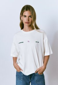 Mujer joven con largo cabello rubio, vistiendo una camiseta blanca de gran tamaño con el texto "UN VISTAZO A NOSOTROS" y una estrella morada, con las manos en los bolsillos de unos vaqueros azules.