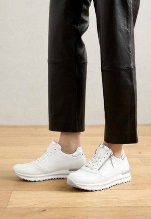Witte sneakers met een gestructureerde bovenkant, zilveren accenten, veters en ritssluiting, gecombineerd met zwarte leren cropped broeken op een houten vloer.