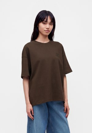 Junge Frau mit glatten schwarzen Haaren trägt ein übergroßes braunes T-Shirt und lockere blaue Jeans vor einem schlichten hellen Hintergrund.