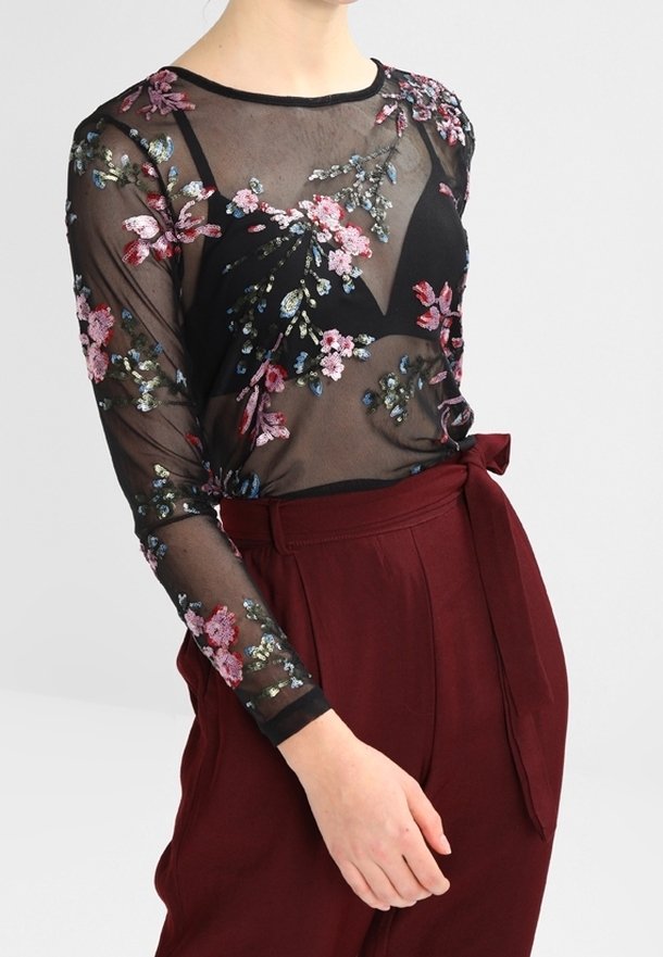 Femme portant un haut noir transparent à manches longues avec une broderie florale colorée sur un soutien-gorge noir, associé à un pantalon bordeaux taille haute avec une ceinture nouée.