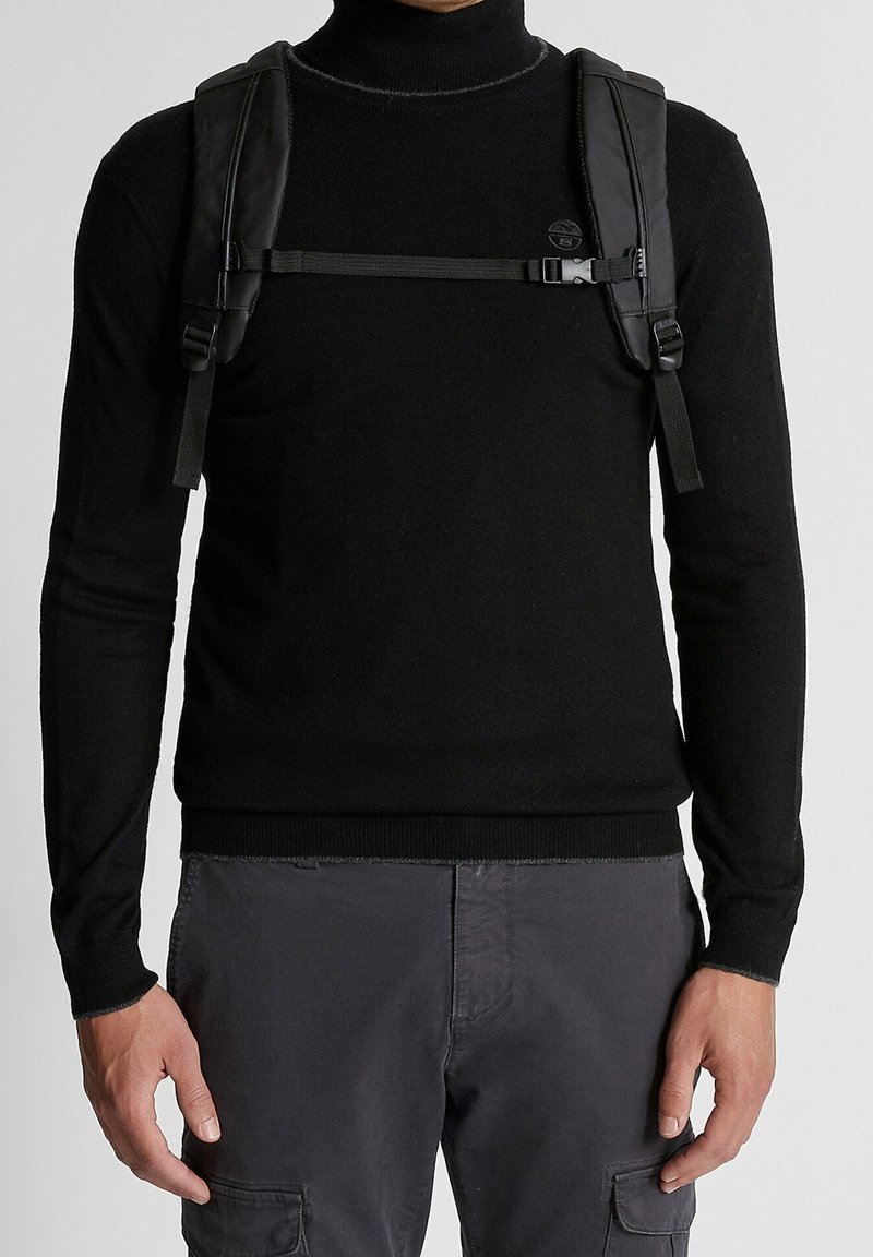 Homme portant un col roulé noir et un pantalon cargo gris, avec des bretelles de sac à dos noir et une boucle de poitrine visibles sur le pull.