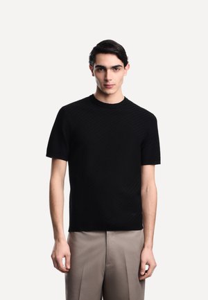 Giovane uomo in piedi, di fronte, che indossa una camicia nera a maniche corte con trama e pantaloni beige chiaro su uno sfondo semplice.