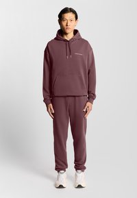 Marron hættetrøje med frontlomme, matchende sweatpants og hvid logo detalje. Har snørebånd, ribbede manchetter og ankellæg.