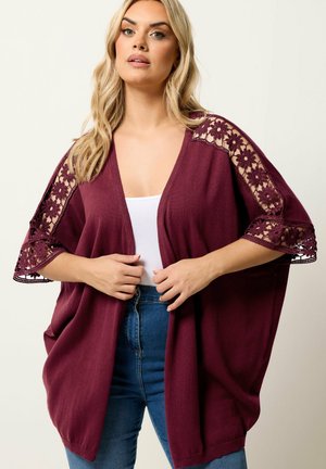 Kvinde iført en burgunder åben cardigan med blomsterblondeudskæringer på ærmerne, hvid top og blå jeans mod en ensfarvet baggrund.