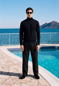 Homme portant un col roulé noir, un pantalon et des lunettes de soleil, debout au bord d'une piscine en pierre avec l'océan et une île rocheuse en arrière-plan.