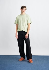 Ung mand står på et blåt tæppe iført en lys grøn t-shirt, løse sorte bukser og brune sko foran en hvid væg.