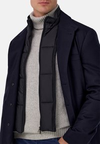 Czarna puchowa kamizelka noszona pod szarym prążkowanym golfem, zestawiona z ciemnogranatową kurtką o dopasowanym, strukturalnym kroju.