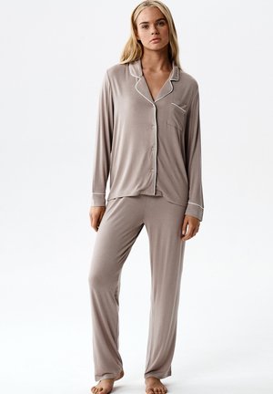 Młoda kobieta ubrana w jasnobrązową, długorękawową, zapinaną na guziki piżamę oraz dopasowane, luźne spodnie piżamowe, stojąca boso na jednolitym białym tle.