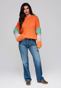 Helles orange gestricktes Sweatshirt mit Ärmeln im mehrfarbigen Muster, kombiniert mit blauen Jeans und braunen Schuhen. Legere Passform und entspanntes Design.