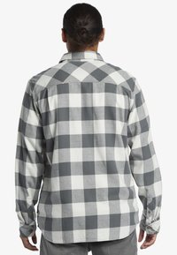 Camicia in flanella a scacchi grigia e bianca con maniche lunghe, colletto a bottoni e finitura liscia e testurizzata. Dettaglio della piega visibile sul retro.