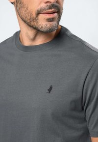 Brodaty mężczyzna w szarej koszulce z okrągłym dekoltem, z małym czarnym haftowanym logo na piersi, na jednolitym jasnym tle.