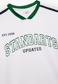 Jersey deportivo blanco con acentos verdes y negros, que presenta "STANDARTS UPDATED" en letras grandes y "EST. 1998" impreso a la izquierda.