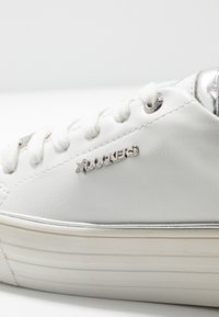 Valkoiset nahkakengät, joissa on pyöreä kärki, teksturoitu pinta, hopeiset yksityiskohdat ja nauhat. Sivulla on "ROCKERS"-logo.