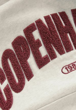 Tissu gris avec une grande broderie en relief de couleur marron, épelant "COPENHAGUE" et l'année "1983" dans un design d'emblème arrondi.