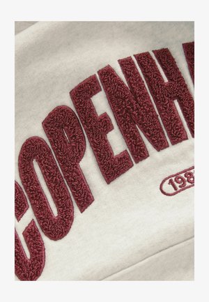 Szara tkanina z dużymi, bordowymi, pluszowo teksturowanymi haftami, na których widnieje napis „COPENHAGENA” oraz rok „1983” w zaokrąglonym projekcie emblematycznym.