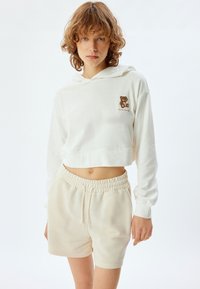 Gekürzter weißer Hoodie mit Bärenstickerei, kombiniert mit beigen Shorts mit elastischem Bund, die eine weiche Baumwollstofftextur zeigen.
