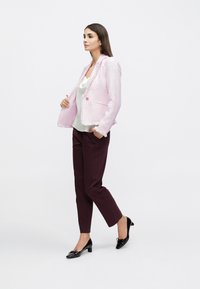 Femme portant un blazer à franges rose clair sur un chemisier blanc, un pantalon violet foncé et des chaussures noires en cuir verni à talons bas, debout devant un fond blanc.