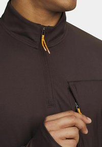 Man in een donkerbruin jack met een kwart ritssluiting en gele ritsen, die de rits van het kleine borstzakje opent.