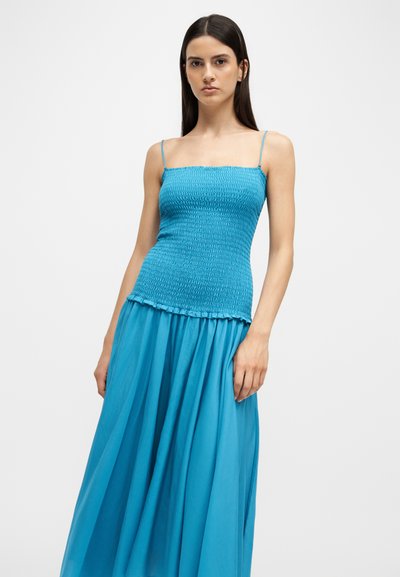 Femme portant un haut bleu texturé sans bretelles et une jupe fluide, debout devant un fond clair uni, faisant face à l'avant.